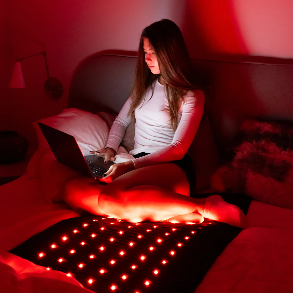 Woman sitting on bed using laptop while on PEMF Far Infrared Heating Mat with red lights.