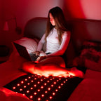 Woman sitting on bed using laptop while on PEMF Far Infrared Heating Mat with red lights.