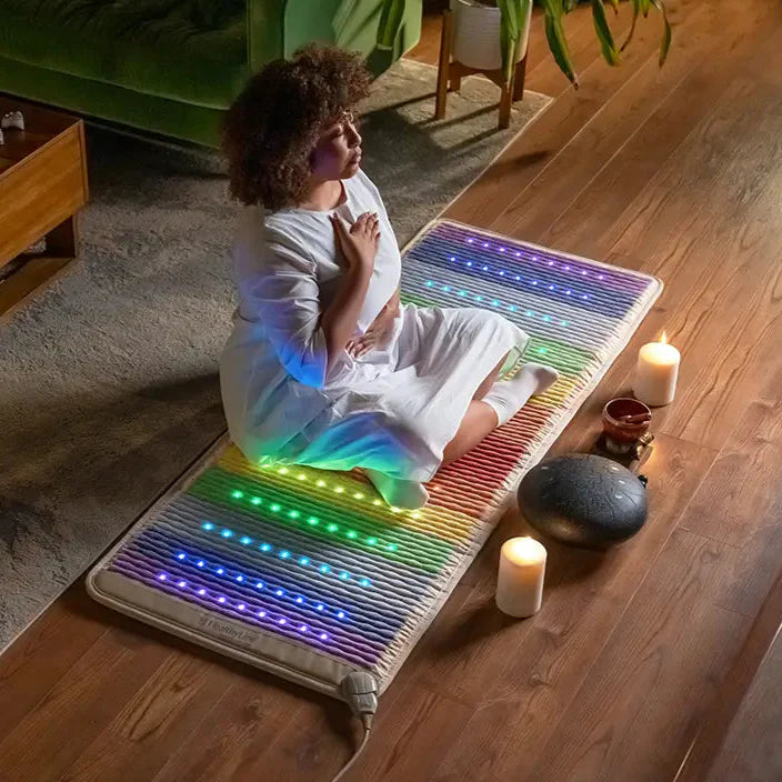 Woman meditating on the Rainbow Chakra PEMF Far Infrared Heating Mat with colorful lights and candles around her.