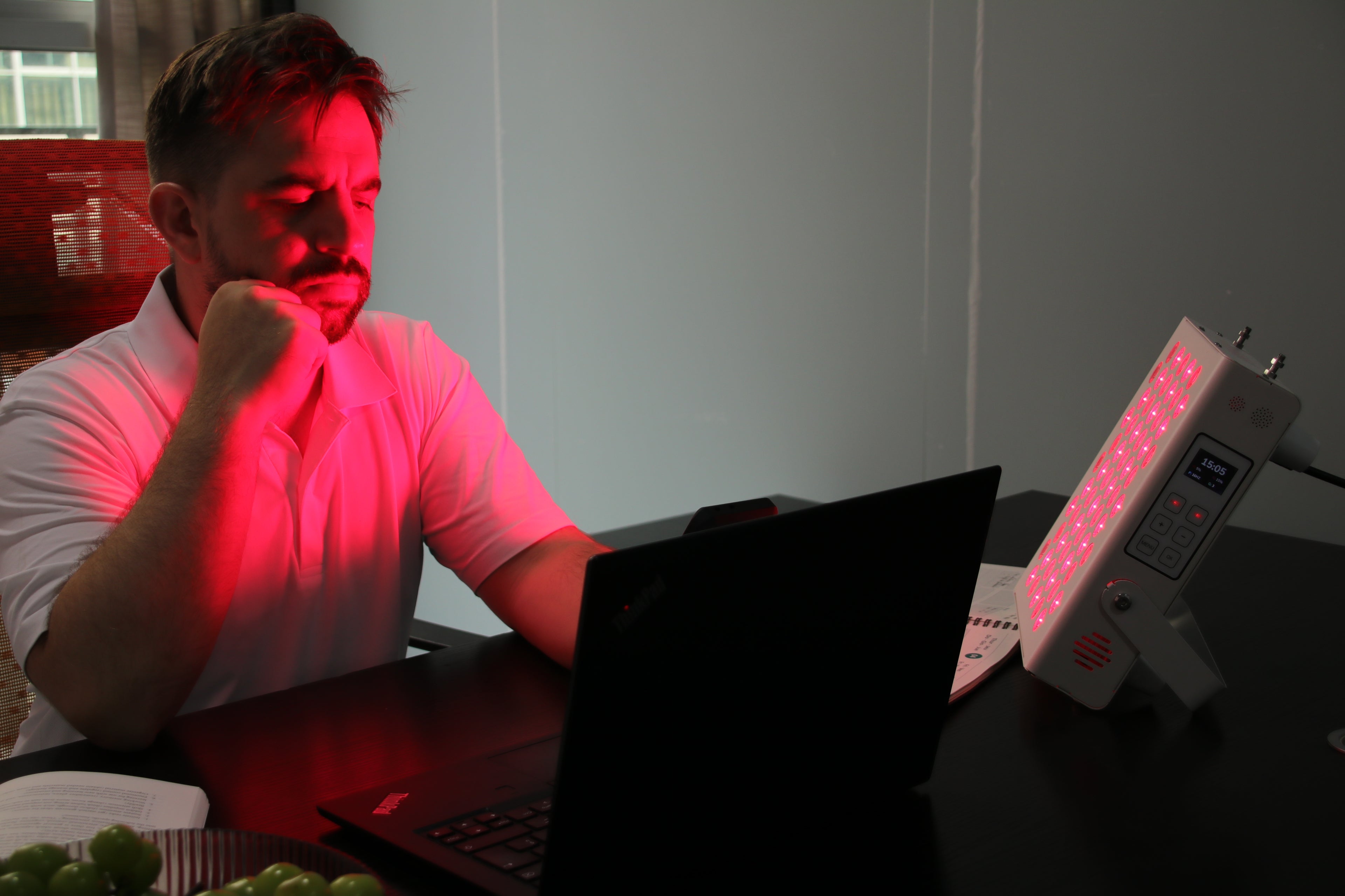 Man using Red Light Therapy Panel while working on a laptop at a desk with a plate of grapes.