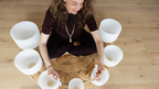 Woman sitting on a woven mat with white sound bowls around her on a wooden floor.